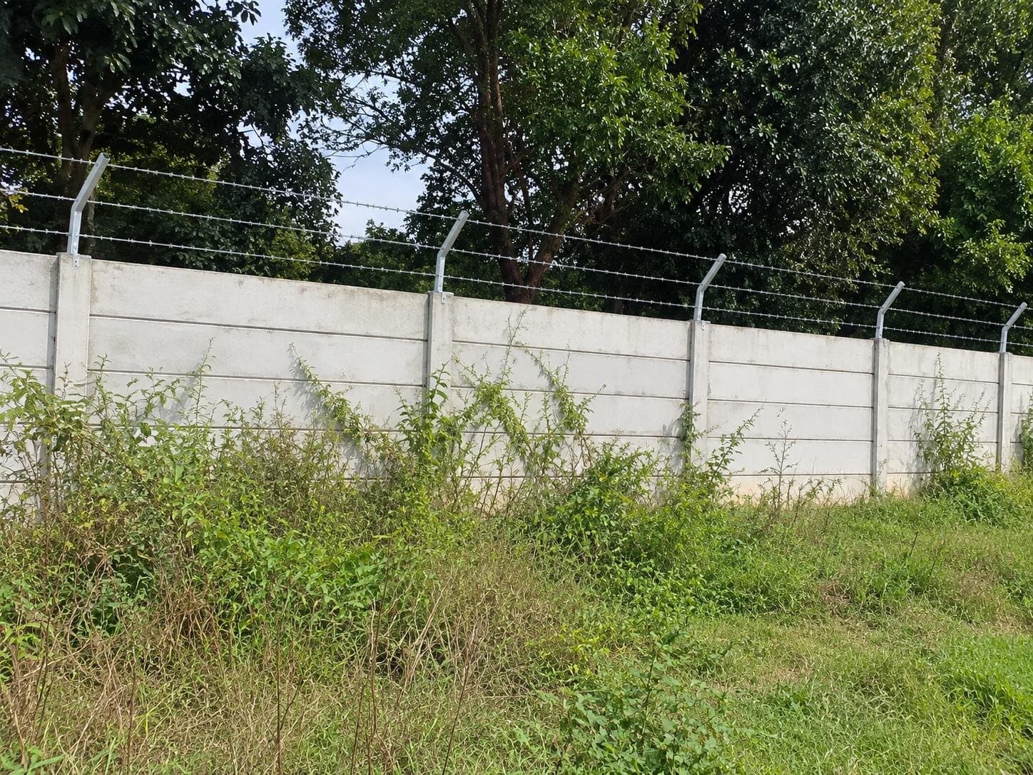 Precast RCC compound wall on site, built from stacked panels between posts, with angled arms carrying barbed wire along the top.